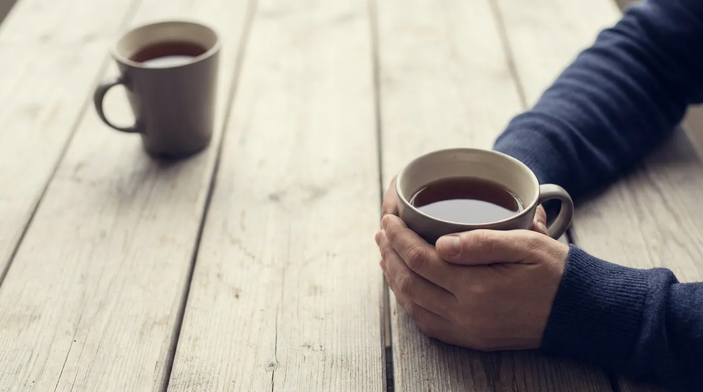 Zwei Tassen auf einem Tisch, zwei Hände halten nur die vordere Tasse, die zweite Tasse steht alleine